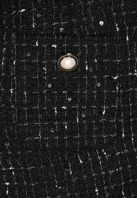 Tejido negro texturizado con acentos en blanco y dorado, que presenta un botón perlado prominente. Patrón de cuadrícula tejido visible en toda la superficie.