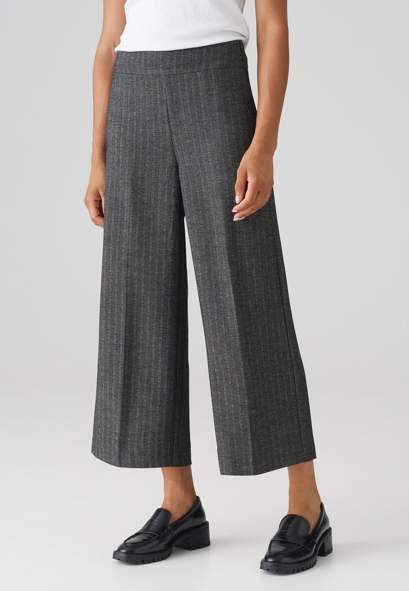 Personne portant un pantalon gris large et court avec un subtil motif en chevrons et des mocassins noirs, debout devant un fond uni.