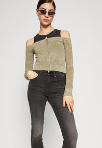 Zeleni maslinasti off-shoulder rebrasti top s patentnim zatvaračem sprijeda, uparen s uskim crnim trapericama. Teksturirana tkanina i dugi rukavi s uskim manžetama.