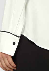 Witte overhemdkraag en manchetten met zwarte piping, voorzien van een zwarte knoop en een gladde stof, gecombineerd met een zwart kledingstuk.