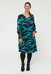 Voľne strihané čierne šaty s tyrkysovým abstraktným vzorom, s výstrihom do V a elastickými dlhými rukávmi. Nosené s čiernymi leggínami a členkovými čižmami.