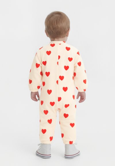 Νήπιο όρθιο με την πλάτη προς τα εμπρός, φορώντας εκρού φορμάκι με κόκκινα μοτίβα καρδιών και ανοιχτό γκρι ψηλά αθλητικά παπούτσια.