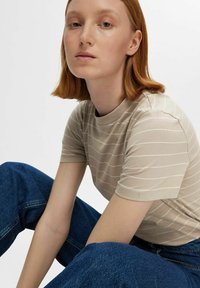 Camiseta de rayas beige con cuello alto, mangas cortas y un diseño ajustado. Lleva puesta unos pantalones vaqueros de mezclilla azules, que muestran una textura suave y un patrón sutil.