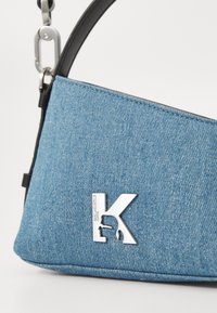 Bolsa de ombro em denim azul claro, com uma alça preta e detalhe do logo prateado. Forma triangular com tecido texturizado e costura limpa.