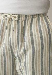 Pantalon en lin rayé beige et gris, avec une taille élastique à cordon de serrage et des poches latérales, mettant en avant un tissu doux et texturé.