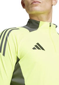 Maglia sportiva gialla a mezzo zip con dettagli grigi, dotata di chiusura a zip e tre strisce nere sulla spalla. Logo ben visibile.
