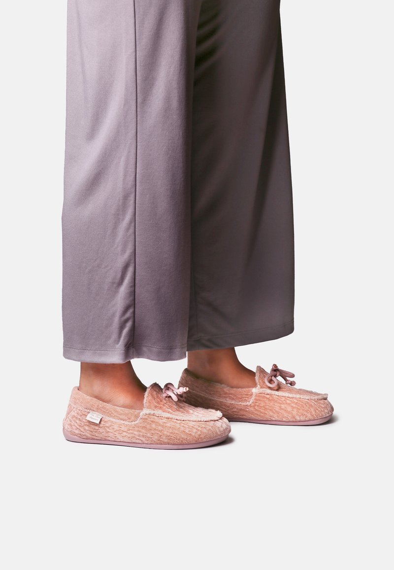 Pieds portant des chaussons mocassins roses doux avec un détail nœud, associés à un pantalon ample lavande, présentés sur fond blanc.