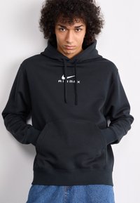 Crna kapuljača od mekog materijala s prednjim džepom, koja sadrži bijeli logo "Air Max" te vezicu, rebraste rukave i rub.