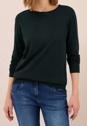 Frau mit schlichtem schwarzem Langarmpullover und blauen Jeans, steht mit entspannt hängenden Händen an den Seiten.