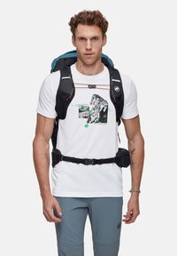 Lätt svart ryggsäck med blå accenter, bärs över en vit t-shirt med en svart och grön bergsgrafik, ihop med grå byxor.