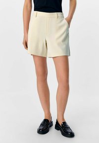 Lichtbeige op maat gemaakte shorts met zijzakken, een gladde textuur en een rechte voorkant. Draagt met zwarte gepolijste instappers.