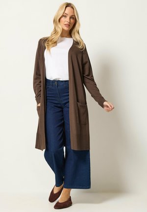 Donna bionda che indossa un lungo cardigan marrone sopra una camicia bianca, jeans blu scuro a gamba larga e scarpe basse marroni, in piedi davanti a uno sfondo semplice.