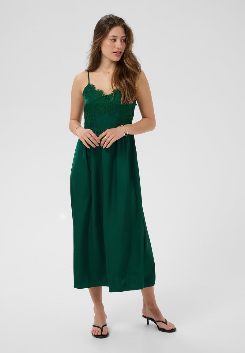 Femme aux longs cheveux ondulés portant une robe midi sans manches vert foncé ornée de dentelle et des sandales à talons noires, debout sur un fond uni.