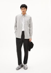 Grijze button-up overhemd met een zak, wit T-shirt eronder, zwarte broek en zilveren sneakers. Houdt een zwarte pet vast. De textuur lijkt glad.