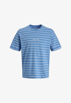 Nastolatek stojący przodem, ubrany w niebieską koszulkę w paski, luźne szare dżinsy oraz brązowe mokasyny na jednolitym białym tle.