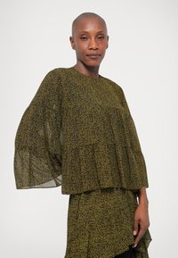 Haut ample de couleur vert foncé avec un motif paisley en jaune. Tissu léger, manches larges et design superposé avec des accents de volants.
