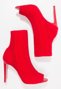 Une paire de bottines en tricot rouges avec des détails perforés, des bouts ouverts et des talons stiletto hauts sur un fond blanc.