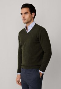 Mörkgrön v-ringad tröja i mjukt material, med ribbade ärmslut och nederkant. Bärs över en ljusblå skjorta med krage.