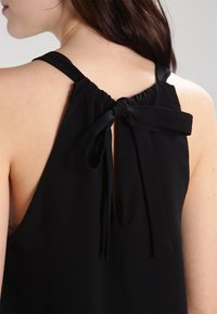 Czarny bezrękawnik z zawiązywaniem z tyłu, z materiałowymi wiązaniami, gładką teksturą i otwartym detalem pleców.