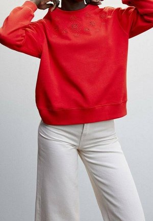 Person trägt leuchtend roten Pullover mit dezentem gesticktem Blumenmuster nahe am Halsausschnitt und offwhite hoch taillierte, weit geschnittene Hose.