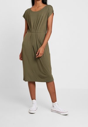 Kvinde iført olivengrøn knælængde kjole med korte ærmer og hvide høje sneakers står foran en ensfarvet lys baggrund.