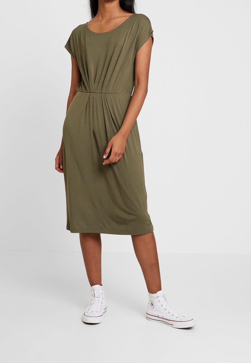 Femme portant une robe vert olive longueur genou à manches courtes et des baskets blanches montantes, debout devant un fond clair uni.