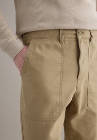 Persona con suéter de punto beige y pantalones marrón claro, con la mano casualmente en el bolsillo delantero, sobre un fondo neutro.