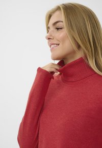 Sarkanais turtlenecks džemperis ar ribotām apkaklēm un aprocēm, izgatavots no mīksta auduma, ar brīvu piegriezumu un gludu tekstūru.