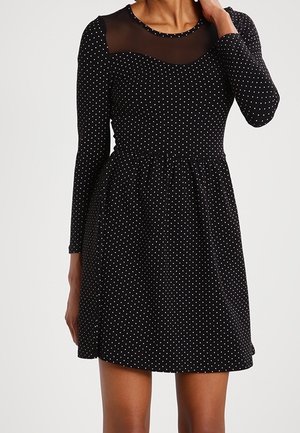 Kobieta w czarnej sukience z długim rękawem w białe kropki, z przezroczystym siateczkowym detalem na wysokości dekoltu.