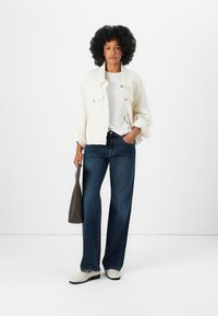 Hvit denimjakke over en hvit skjorte, kombinert med mørkeblå vide jeans og kremfargede sko. En grå håndveske holdes i venstre hånd.