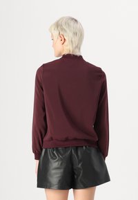 Bordeaux genser med høy krage og ribbestrikkede mansjetter, sammen med blanke svarte shorts i kunstlær. Glatt tekstur, avslappet passform.