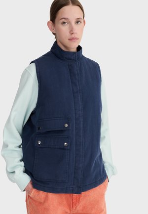 Junge Person trägt eine marineblaue ärmellose Weste mit geknöpften Taschen über einem hellblauen Langarmhemd und orangenen Hosen.