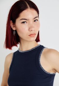 Top azul marinho canelado sem mangas com realces de costura branca; design ajustado revela os ombros. Modelo usa brincos de argola prateados.