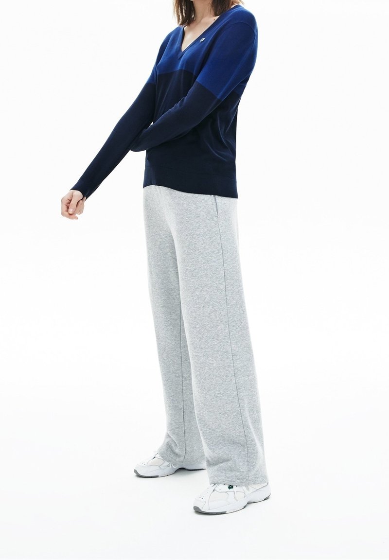 Pull à col en V bleu marine et bleu en matière douce, associé à un pantalon de survêtement large gris clair avec coutures latérales, et des baskets blanches.