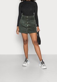 Mini-jupe en velours côtelé vert avec boutons sur le devant, deux poches latérales et une finition texturée, portée avec un top noir ajusté à manches longues et des baskets blanches.