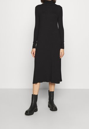 Femme portant une robe midi noire à manches longues et col roulé, associée à des bottines noires, debout devant un fond uni.
