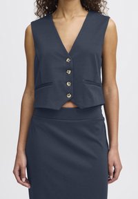 Nainen, jolla on päällään tummansininen hihaton napillinen liivi ja siihen sopiva korkeavyötäröinen hame, seisoo yksinkertaista vaaleaa taustaa vasten.