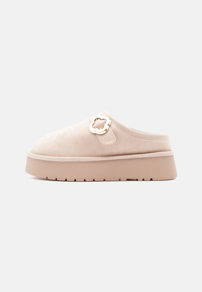 Beige mockasuinsko slip-on-sko med en kraftig ljusbeige sula, försedd med en dekorativ gyllene blommadetalj på sidan. Texturerad ovandel och slät yta.