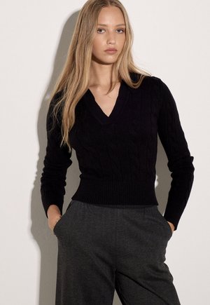 Jeune femme aux longs cheveux blonds porte un pull noir à col en V et un pantalon gris foncé, mains dans les poches, sur un fond uni.