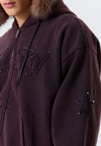 Nærbillede af person iført mørkeblå hoodie med pelskrave, broderet tekst på brystet og stjernemærkat på ærmet.