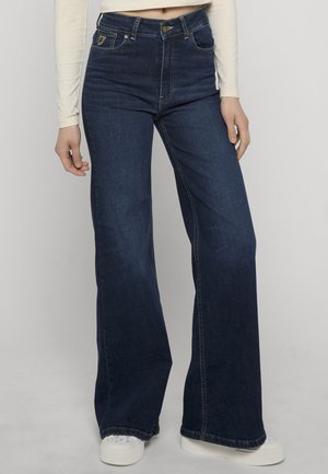 Vrouw draagt donkerblauwe flared jeans met hoge taille en voorzakken, gecombineerd met witte platform sneakers en een beige top met lange mouwen.