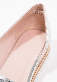 Chaussure à enfiler en métal argenté avec une doublure intérieure lisse, de forme arrondie, avec une texture subtile et une semelle beige.