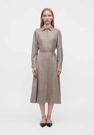Woman standing facing forward wearing a long-sleeve beige buttoned dress with a waist belt and black pointed shoes.