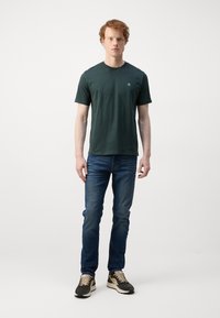 Dark green crew-neck t-shirt, cotton material, short sleeves, featuring a small logo on the left chest. Paired with blue jeans and sneakers.
