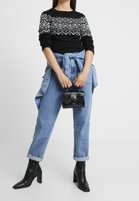 Schwarzer Pullover mit weißen geometrischen Mustern, helle Jeans aus Denim, blaues Hemd, das in der Taille gebunden ist, und eine kleine schwarze Handtasche mit Struktur. Schwarze Stiefel mit Absatz.