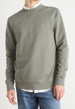 Mężczyzna w oliwkowej bluzy crewneck noszący pod białą koszulą i niebieskimi dżinsami, z naszywką logo "BOSS" na piersi.