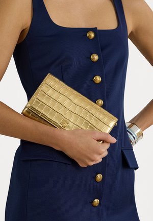 Gold croc-embossed clutch with a sleek rectangular shape, held against a navy textured dress featuring gold buttons.