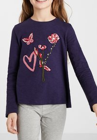 Dítě oblečené v námořnické dlouhé tričko s růžovým srdcem, červenými a růžovými květinami a vyšitými motýlky, spárované se světlými šedými kalhotami.