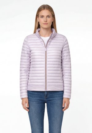 Kobieta w jasnofioletowej pikowanej kurtce na białej koszuli i niebieskich dżinsach, stojąca na tle jednolitego białego tła.