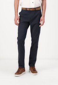 Tamno plave pamučne hlače slim fit kroja, uparene s smešnim kožnim remenom i smešnim tenisicama. Glatka tekstura, bez vidljivih uzoraka.